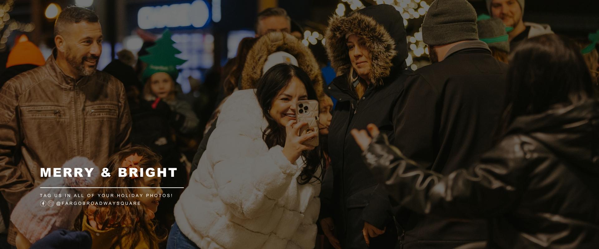 This image shows people taking holiday photos in front the christmas tree in downtown fargo at broadway square with text saying tag us in all of your holiday photos