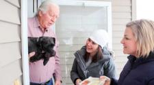 Photo shows volunteers delivering Meals on Wheels to senior holding a dog.