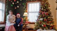 Santa and Mrs. Claus sitting in house on Santa's chair next to lamp and Christmas Tree decorated 