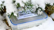 A plastic box with a label on the lid "Official Geocaching" under a snow covered pine tree