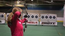 View from behind of a lady in a pink shirt shooting a bow and arrow at a target inside of a building