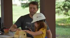 A Father and Daughter sitting at a picnic table putting together a wooden birdhouse.  The daughter is in a hard hat and yellow shirt. 