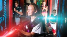a girl stands in running stance smiling playing laser tag with a red flashing laser tag gun in her hands wearing a laser tag vest. Behind her stands two boys and a girl ready to play