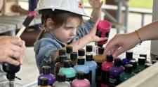 Young girl picks out the perfect paint color at a counter with 20+ paint bottles for her wood bird house, wearing a construction hard hat.