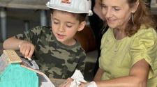 A woman sits with a young boy at a picnic table while they paint their bird house.  The boy is wearing a hard hat. 