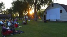 View of the Rheault Farm barn with the large white movie screen attached with the sun setting behind the trees and playground equipment while people relax on the lawn waiting for the movie to start
