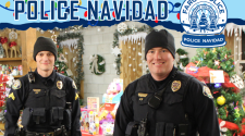 Two Policemen standing in the Santa Village barn with loads of donated gifts and food. POLICE NAVIDAD above the men and the Fargo Police snow globe