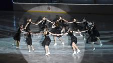 11 older teens skating in a circle in black dresses around a girl in the middle skating in a spin, ice skating to a routine for the Fargo Parks Ice Revue