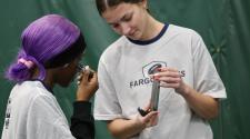 Two high school girls at an indoor field, one using eye black under her eyes while the other holds a phone for a mirror