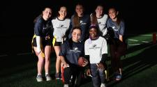Group of high school girls wearing Fargo Parks flag football shirts pose together on an indoor turf field, smiling and holding a football.