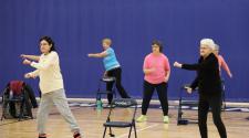 Group of older adults doing balance exercises in a gym with chairs to help maintain balance. 