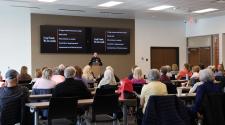 A police officer presents fraud prevention information to a group of older adults seated in a community meeting room.