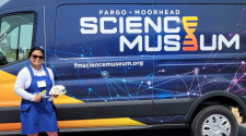 Woman standing in front of a large Fargo Moorhead Science Museum wrapped cargo van holding a beaver skull and magnifying glass