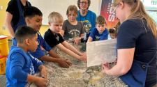 A group of kids standing around a mock river bed with sand and water running through learning from a staff member about dam erosion. 