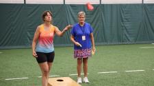 Two women play bean bag toss indoors on a turf field during the North Dakota Senior Games.