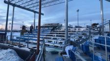 Workers install and repair aluminum bleachers at an outdoor sports complex during winter, with snow on the ground and stadium light poles in the background.
