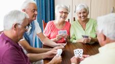 A group of older adults gathered around a table playing cards together. Several participants hold cards while smiling and socializing.