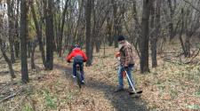 A cyclist rides along a narrow wooded trail while a park worker uses a string trimmer to maintain the trail. Fallen leaves cover the surrounding forest floor.