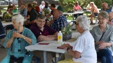Older adults sit around picnic tables under a park shelter, smiling and talking together during an outdoor social gathering.